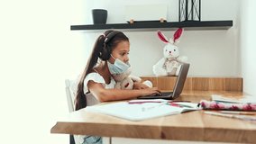 Little girl wearing headphones and a protective face mask while attending an online school lesson, using her laptop and writing in her notebook at a desk during the coronavirus pandemic - Powered by Shutterstock - Get 15% off with code: PIKWIZARD15