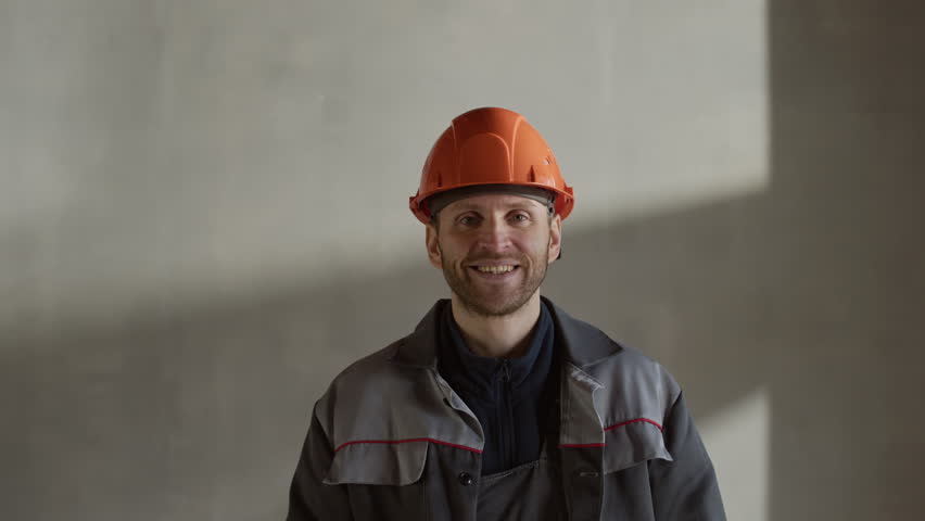 Zoom-in chest up slowmo portrait of Caucasian construction worker in protective workwear and hard hat smiling kindly at camera while standing inside unfinished building