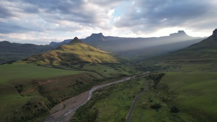Aerial drone footage of uKhahlamba Drakensberg Mountains in KwaZulu Natal South Africa showing winding river through green valley illuminated by sunlight and dramatic mountain peaks under cloudy sky