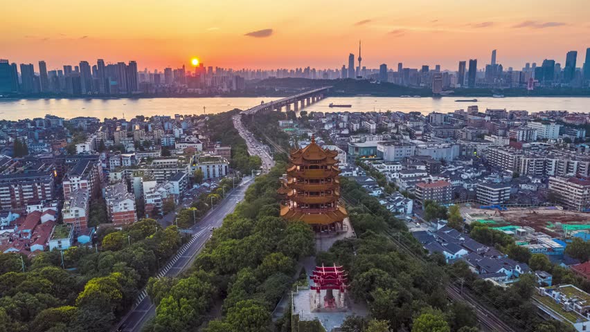 Aerial view of Yellow Crane Tower landmark in Wuhan, China at sunset with urban skyline, Yangtze River bridge and traditional Chinese architecture surrounded by greenery.