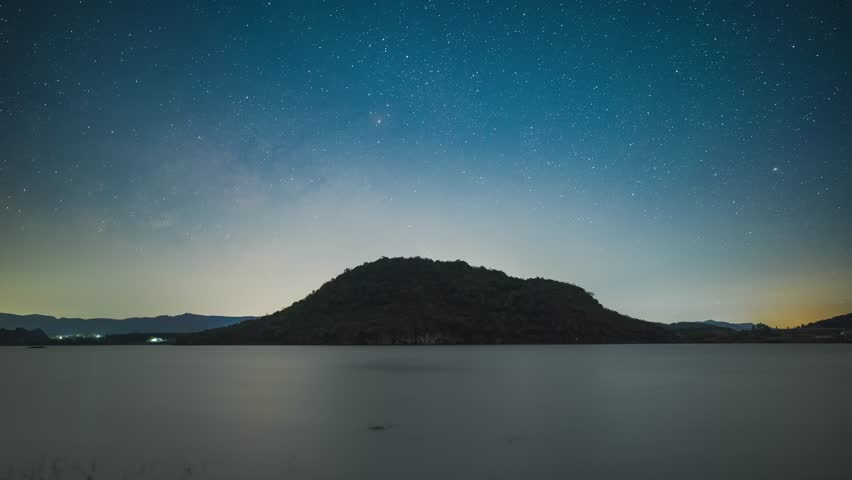 Stunning night landscape of Haifeng wetland in Yunnan, China with starry sky and Milky Way over serene lake and silhouetted mountain under twilight blue hour sky.