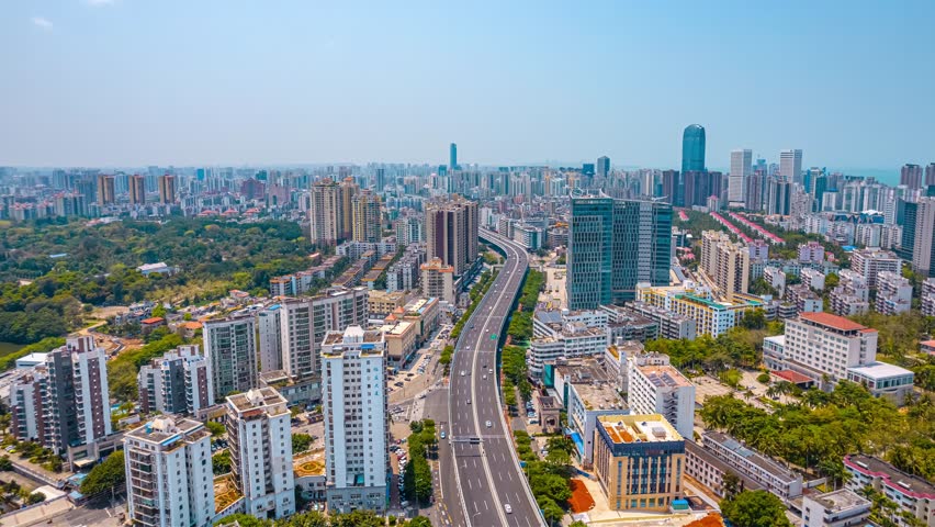 Aerial time lapse view of Longhua District in Haikou, Hainan Island, showcasing modern urban development with highways, skyscrapers and residential buildings under blue sky.