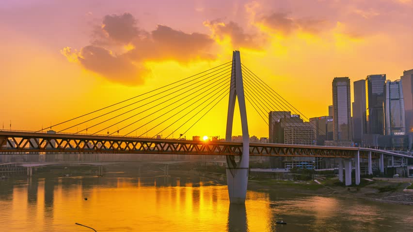 Stunning sunset view of Qianliamen Bridge in Chongqing, China with golden sky reflections on water and modern city skyline silhouetted against dramatic orange clouds during dusk.