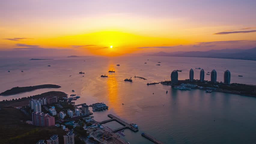 Stunning aerial view of Sanya, China at sunset with golden sky reflecting on calm sea waters, featuring Phoenix Island and coastal buildings in beautiful evening light