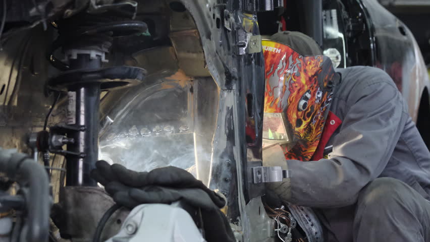 Worker in protective mask welds carcass of damaged car in repair shop. Mechanic fixes vehicle body with welding machine at service station