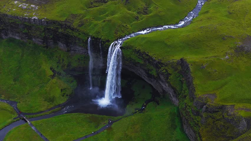 Aerial drone view shows Seljalandsfoss on Iceland's south coast as water plunges over basalt, spray fans out, paths and a footbridge weave, visitors appear small under soft light.