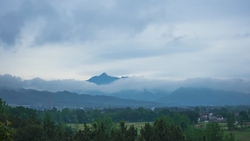 Beautiful mountain landscape with layered peaks emerging through clouds and mist, featuring lush green forests and rural valley below in atmospheric natural scenery.