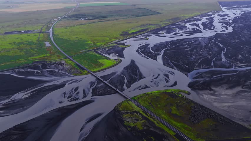 Aerial view shows a straight road and bridge across braided glacial channels on black volcanic outwash plains in south Iceland, with mossy islands in soft daylight.