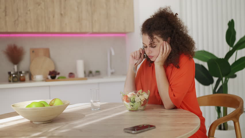 Sad young woman eating salad without appetite, struggling with strict diet