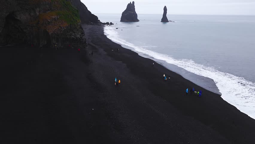 High aerial view of Reynisfjara near Vik, Iceland. Reynisdrangar sea stacks rise offshore, basalt cliffs and cave line the beach as waves roll and visitors walk.