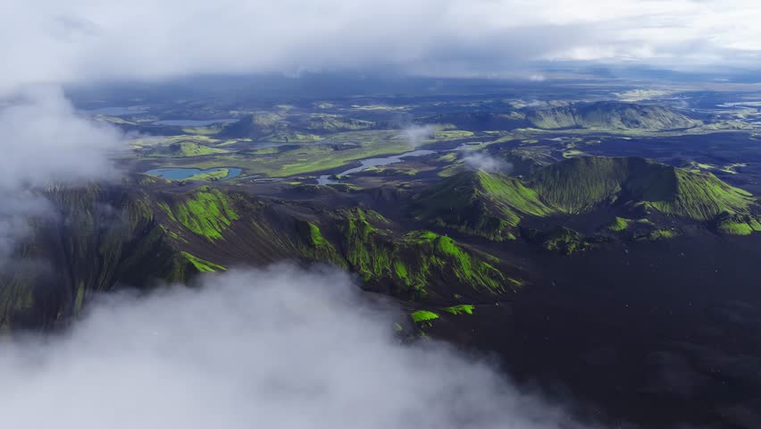 Aerial view of Icelandic Highlands with moss green craters, turquoise lakes, and braided rivers under soft overcast light. Slow cinematic movement reveals ridges.