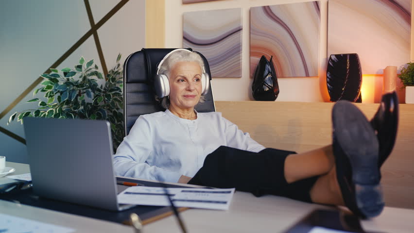 Restful senior woman in headset listening to music, pleased with finished work