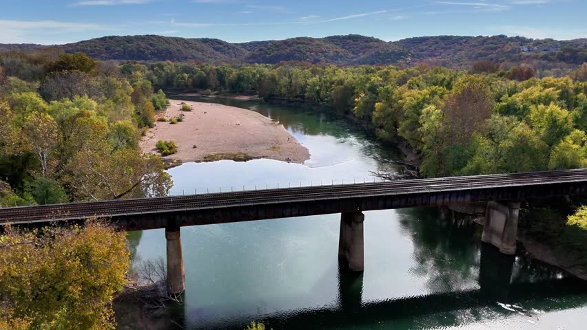 Aerial view of a rail bridge crossing the serene Meramec River, framed by autumn trees and sandy shores under a bright sky, Ballwin, Missouri, United States.