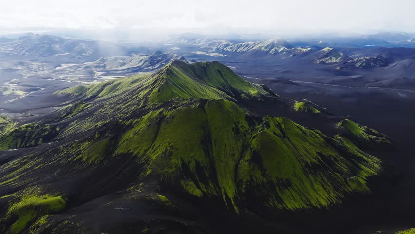 Aerial view shows moss covered cones, cratered ridges, and black ash plains in Icelandic highlands. Camera tracks and pans in soft daylight with wide framing.