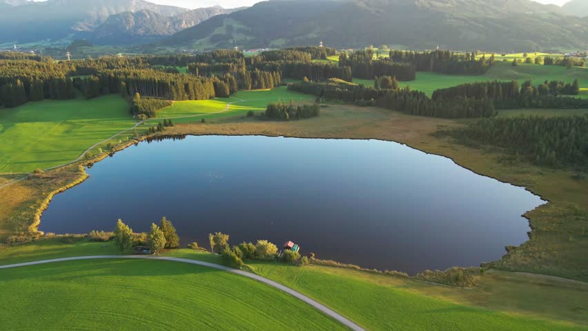 Aerial view of a serene lake reflecting the sky, surrounded by lush green meadows and dense forests, with mountain ranges in the backdrop, Nesselwang, Bayern, Germany.
