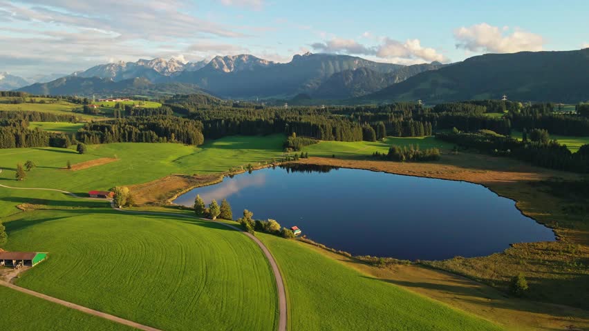 Aerial view of a serene lake reflecting the sky, surrounded by green fields, forests, and distant mountains, creating a tranquil landscape, Nesselwang, Bayern, Germany.