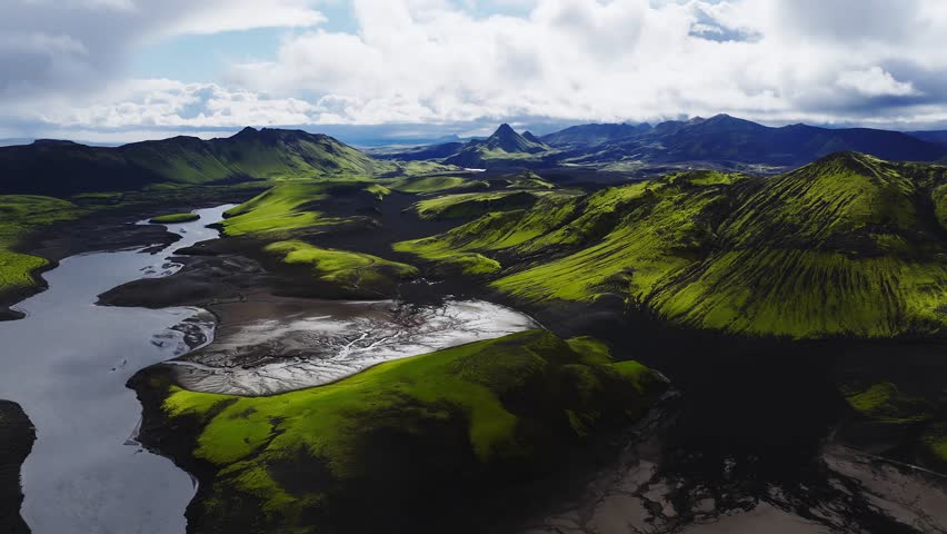 Aerial view shows moss clad cones, black sand flats, and braided glacial river in Iceland highlands, with crater like hills and low clouds drifting across the valley.