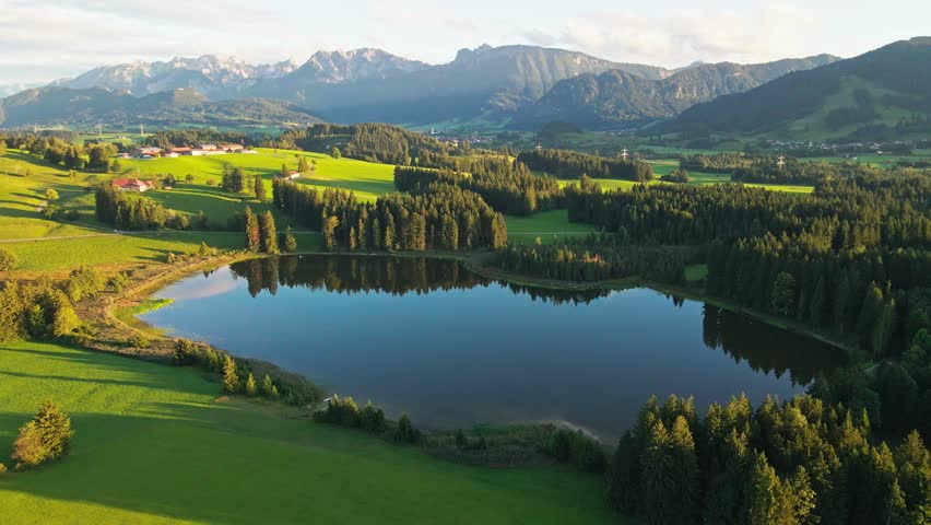 Aerial view of a picturesque lake surrounded by green fields, forests, and mountains offering a serene landscape, Nesselwang, Bayern, Germany.