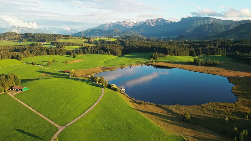 Aerial view of a serene lake mirroring the sky, surrounded by vibrant green fields and forests, creating a picturesque landscape, Nesselwang, Bayern, Germany.