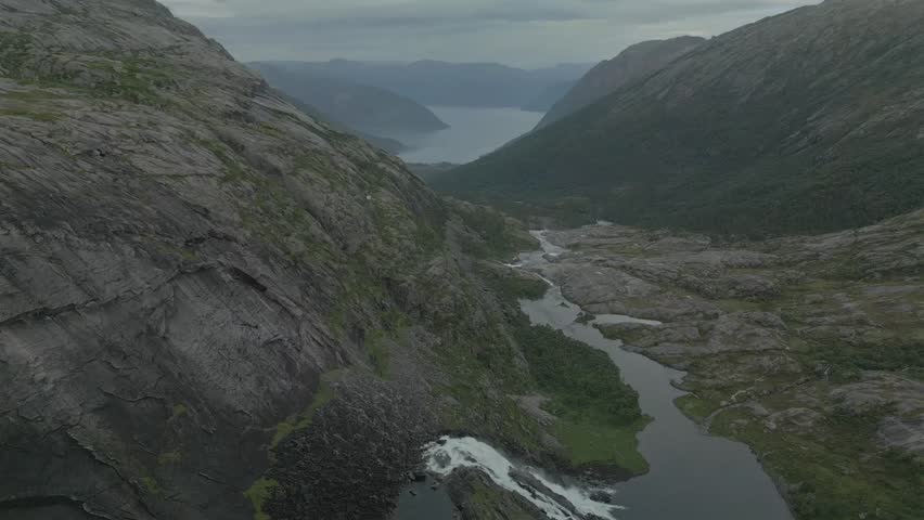 Aerial view of a river flowing through rocky terrain between mountains, a cascade and a distant fjord, Kinsarvik, Vestland, Norway.