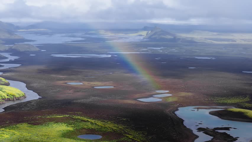 Aerial view pans over moss ridges, dark lava plain, small ponds and rivers in Iceland. Low clouds and light move as a rainbow touches the plain and lakes reflect.