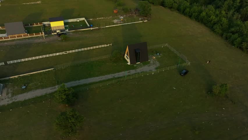 A single triangular cabin stands alone in a vast open field at sunset, surrounded by long paths and soft hills fading into the evening light.