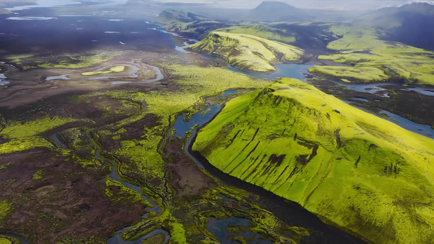 Aerial view in Iceland shows chartreuse slopes, dark lava plains, braided rivers, turquoise lakes, and a faint rainbow under soft daylight near Landmannalaugar.