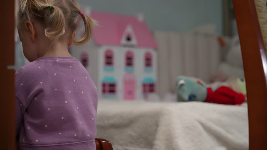 Blonde toddler with pigtails plays intently with her dollhouse and toys in cozy room, sharing heartwarming moments of contemplation and observation.