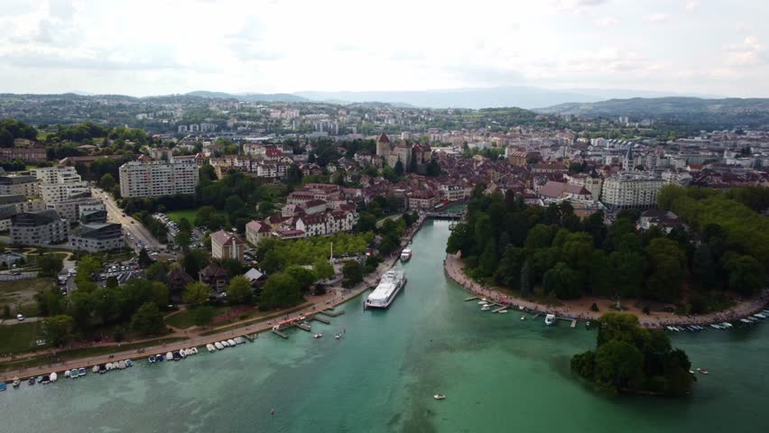 Aerial footage over Annecy in Southern France. Footage moves sideways to the right over the ferry port to a park. Views showing the city with buildings stretching into the distance. Boats moored.