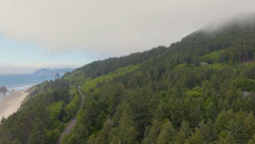 Cinematic aerial tilt down over trees and road along Arcadia Beach on the Oregon Coast with Cannon Beach on the horizon as a red car drives up the highway on a clear beautiful day.