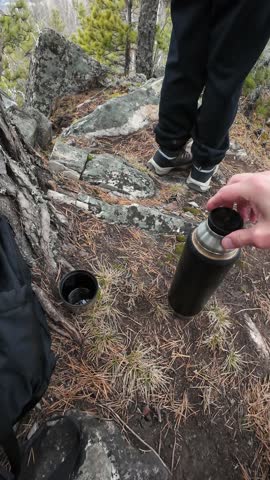 Companion offers thermos at granite lookout while another stands at ridge, pouring into cup near tree roots, hiking boots and backpack visible, shared pause and panoramic valley view