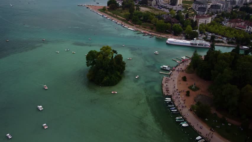 Aerial footage panning up over a suburban area of Annecy in France. Footage reveals boats moored on the shore and a building complex with hills in the background.