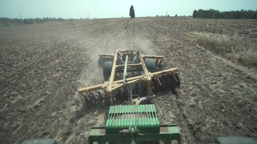 A disc harrow plows a dry field under bright daylight, raising dust clouds. Blue summer skies. Field ready for sowing. Shot from tractor driver POV. Modern agriculture in Israel, the Holy land