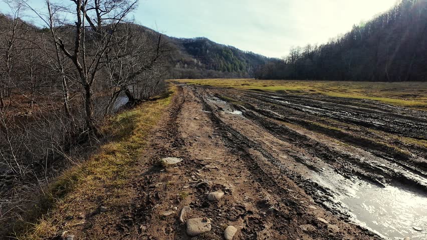 Muddy dirt road through valley with bare tree, rutted tracks and puddles near slow river under cold sky; late autumn field, distant hills and worn path create contemplative rural mood