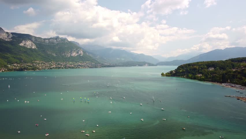 Vast open views of lake Annecy. Drone footage moves backwards over the lake with many boats and yacht on the water and with stunning mountains in the background.