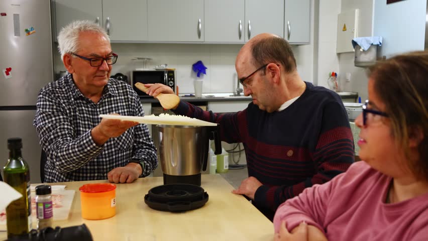 Group of people with intellectual disabilities learning to cook with a caregiver in a kitchen. Man in a wheelchair adding ingredients to a food processor