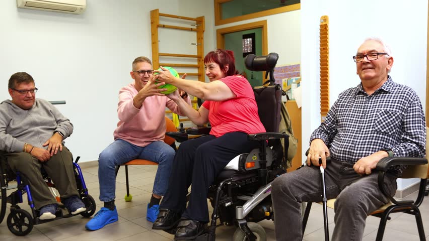 Adults with disabilities in wheelchairs happily participating in a group physical therapy session. The therapist engages them by passing a ball