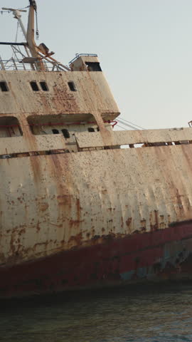 A vertical shot of a weathered shipwreck resting on a rocky coastline, with waves gently lapping against the hull.