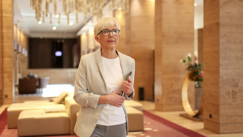 Confident senior businesswoman wearing glasses striding through a sleek hotel lobby. This sophisticated and accomplished female entrepreneur is on a successful business trip