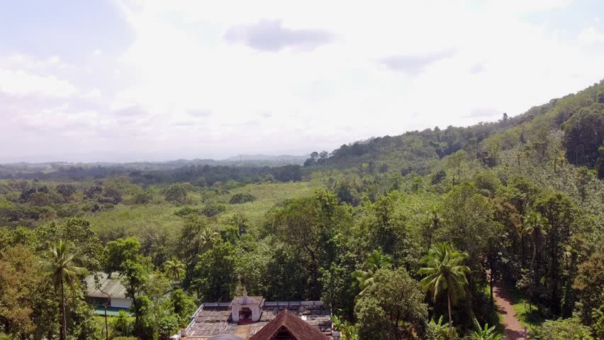 Cinematic aerial descent revealing a historic, white Christian church with traditional tiled roofs, a bell tower, and a lush forested hillside backdrop in Kerala, India.
