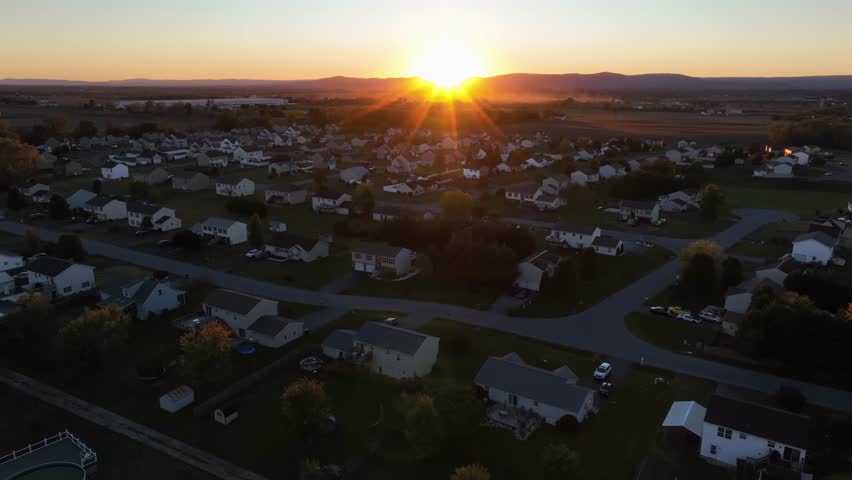 Aerial establishing shot of peaceful suburb neighborhood in america at golden sunrise in the morning. Multi-colored trees in fall season. Single family houses with white facade and grey roofs.