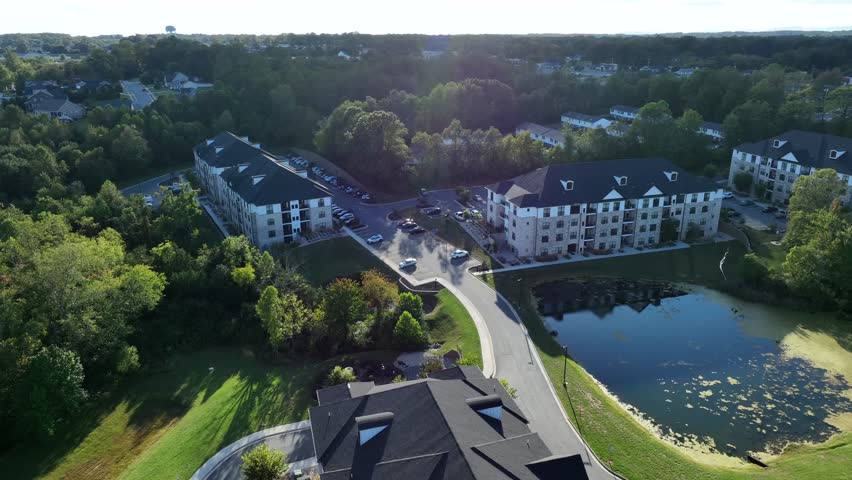 Slow drone flight over street along private lake in luxury suburb neighborhood with multi-family units and apartment complex of USA. Aerial approaching shot. Autumn season with colored trees and sun.