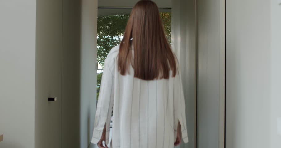 A woman walks down a hallway and opens a door to the outside, revealing a view of trees. She is wearing a striped, loose-fitting shirt. This moment evokes a sense of peace and tranquility.