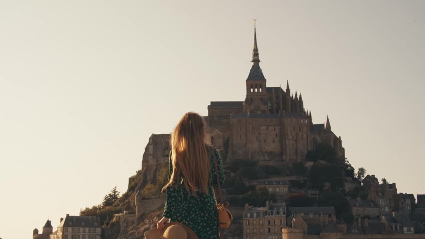happy young woman tourist traveler in hat walking towards to saint michel castle, hair blowing in wind. Abbey Mont Saint-Michel Normandy France. view from outside fortress city. Abbey in background.