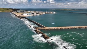 Aerial footage of Helgoland’s harbor with breakwater cutting into North Sea, waves crash along seawall as boats lie moored inside sheltered marina, low-lying island of Düne and red sandstone cliffs. - Powered by Shutterstock - Get 15% off with code: PIKWIZARD15