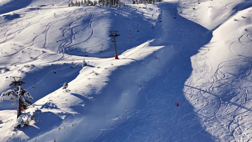 Aerial footage of snowy mountain slopes showing chairlifts carrying riders and people skiing or snowboarding on the piste, set in a bright alpine winter landscape.