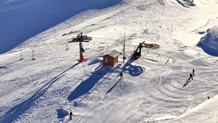 Aerial footage of winter sports riders unloading from a chairlift at the mountain summit, with snowy slopes, lift machinery, and wide alpine terrain in view.