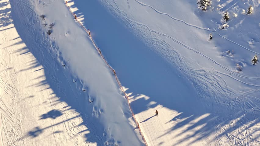 Aerial footage of skiers and snowboarders gliding down a snowy mountain slope, showcasing smooth winter sports action on a wide, sunlit piste.