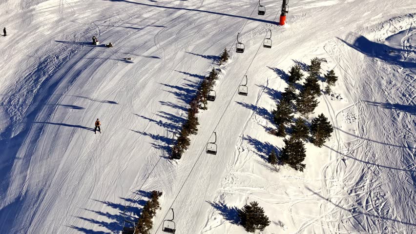 Aerial footage of snowy mountain slopes showing chairlifts carrying riders and people skiing or snowboarding on the piste, set in a bright alpine winter landscape.