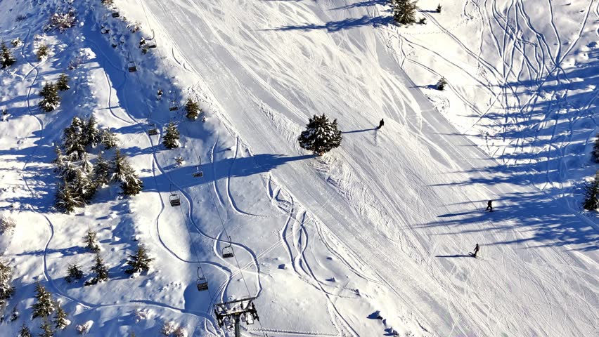 Aerial footage of skiers and snowboarders gliding down a snowy mountain slope, showcasing smooth winter sports action on a wide, sunlit piste.